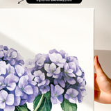 a hand holding a white canvas print of a painting of purple hydrangea flowers.