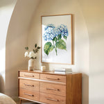a wooden dresser with a framed painting of blue hydrangeas hanging on the wall above it. The dresser has six drawers and a vase of white flowers on top.