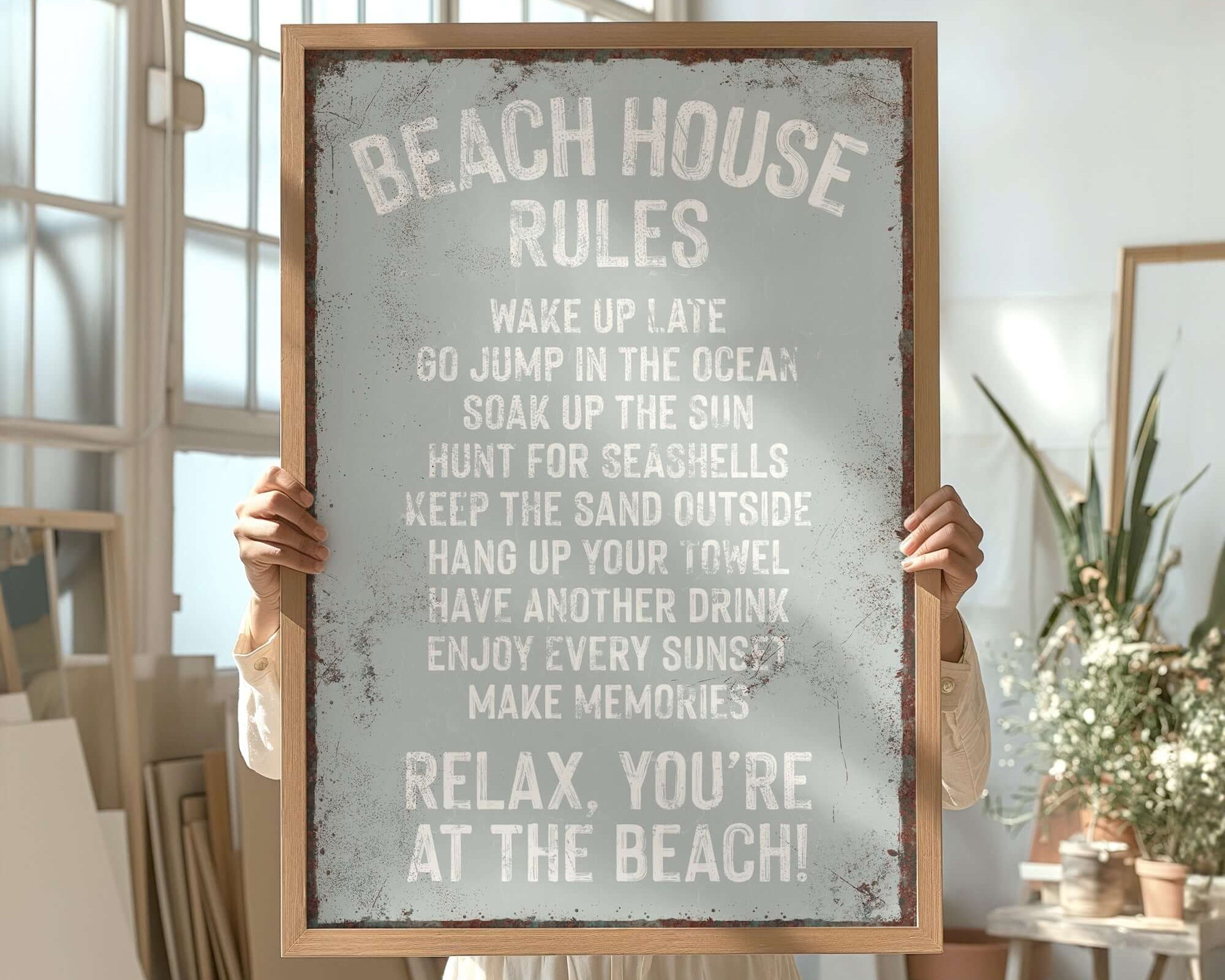 Person holding a Beach House Rules Sign (Rusted Edge, Smoke) in a sunlit room with large windows, houseplants, and modern farmhouse decor.