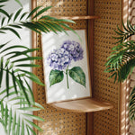 a framed painting of a purple hydrangea flower on a wooden shelf, surrounded by lush green leaves.