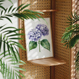 a framed painting of a purple hydrangea flower on a wooden shelf, surrounded by lush green leaves.