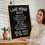 A woman is holding a chalkboard sign with a list of rules for a lake house, including instructions for guests to wake up late, jump in the lake, take a nap, drink, and enjoy the sunset.
