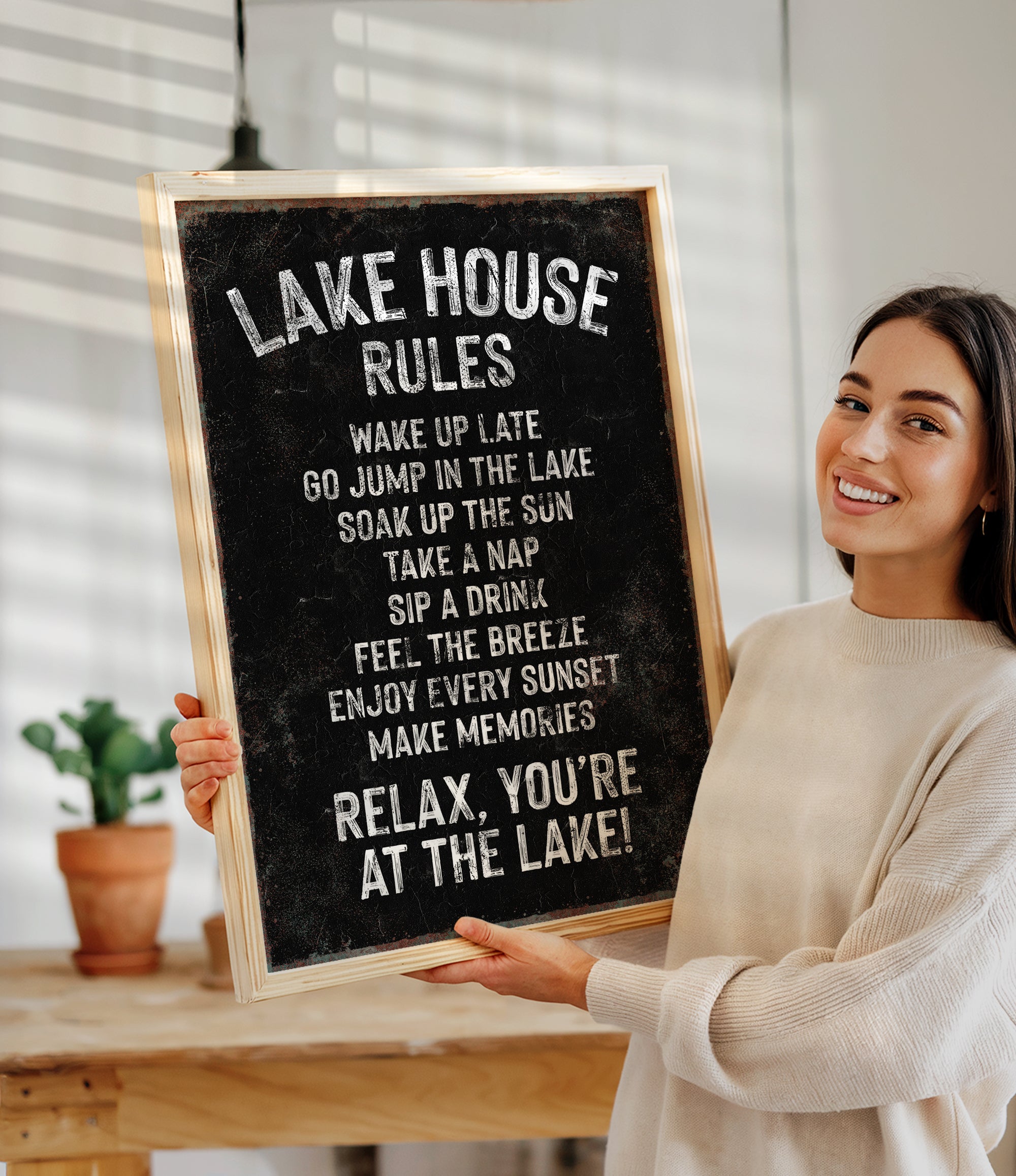 A woman is holding a chalkboard sign with a list of rules for a lake house, including instructions for guests to wake up late, jump in the lake, take a nap, drink, and enjoy the sunset.