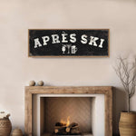a cozy fireplace with a rustic wooden mantel, a black sign with white text that reads "APRES SKI", and a wicker basket on the floor next to the fireplace.