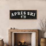 a cozy fireplace with a rustic wooden mantel, a black sign with white text that reads "APRES SKI", and a wicker basket on the floor next to the fireplace.