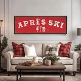 a cozy living room with a large red sign that says "APRÈS SKI" in bold white letters. The sign is mounted on a white wall above a beige sofa with red and white plaid throw pillows. In front of the sofa, there is a wooden coffee table with a tray and a mug. The room also includes a potted plant, a lamp, and a vase, creating a warm and inviting atmosphere.