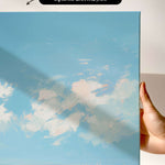 a hand holding a framed canvas print of a serene landscape depicting a blue sky with fluffy white clouds and a body of water with sailboats.