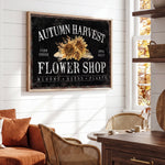 a rustic-style living room with a large framed sign that reads "Autumn Harvest Flower Shop" above a wooden table. The sign has a sunflower design and the text "FARM FRESH FLOWER SHOP BLOOMS SEEDS PLANTS" written on it. The room has a cozy atmosphere with a couch, chairs, and a wooden cabinet.
