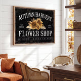 a rustic-style living room with a large framed sign that reads "Autumn Harvest Flower Shop" above a wooden table. The sign has a sunflower design and the text "FARM FRESH FLOWER SHOP BLOOMS SEEDS PLANTS" written on it. The room has a cozy atmosphere with a couch, chairs, and a wooden cabinet.