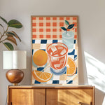 a wooden cabinet with a lamp on top, and a framed artwork depicting a glass of orange juice and slices of orange on a checkered tablecloth.