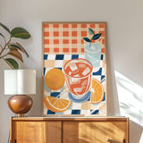a wooden cabinet with a lamp on top, and a framed artwork depicting a glass of orange juice and slices of orange on a checkered tablecloth.