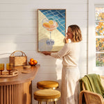 A woman hangs a framed artwork on a white wall in a cozy kitchen setting.