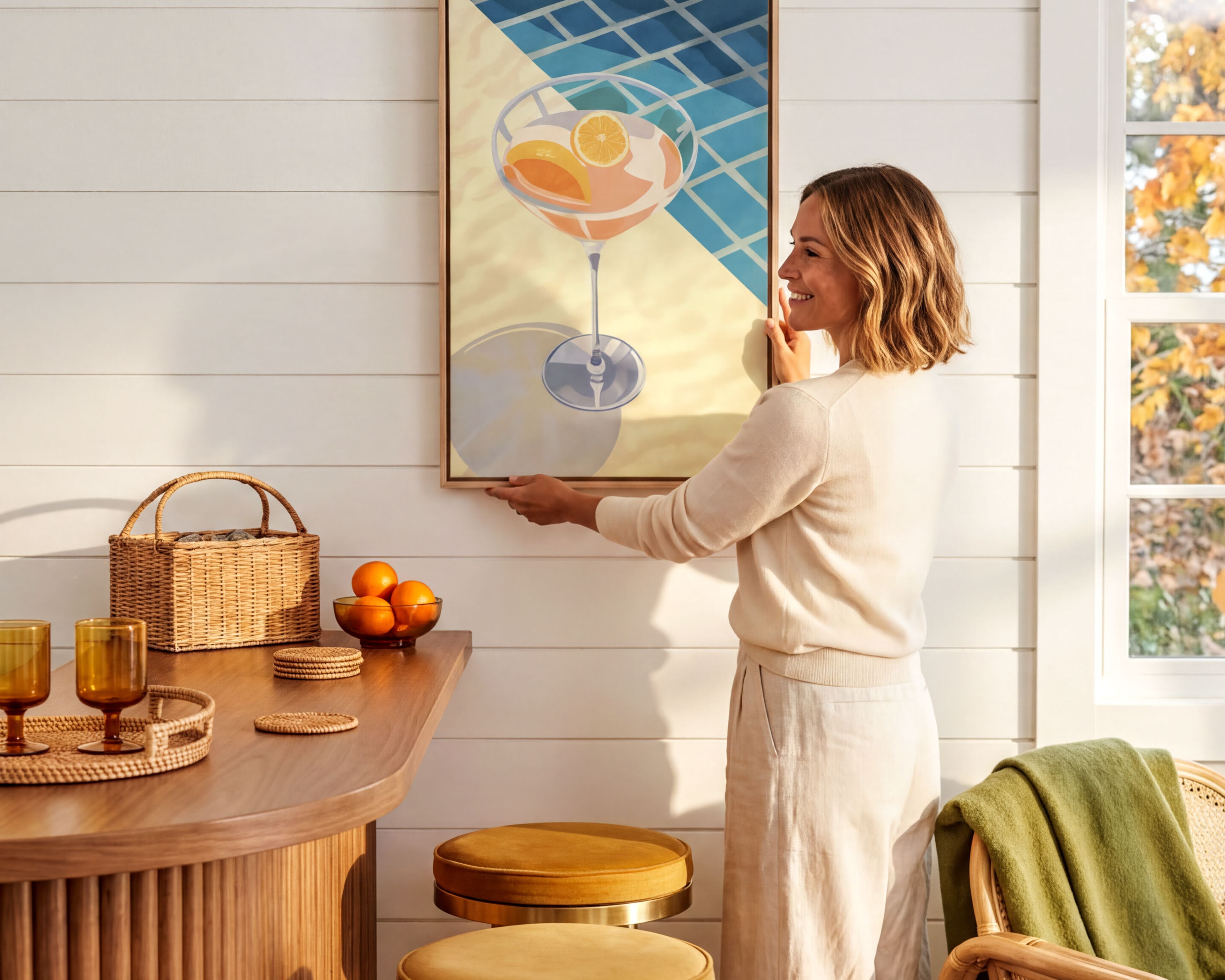 A woman hangs a framed artwork on a white wall in a cozy kitchen setting.