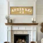 a cozy living room with a fireplace adorned with a decorative tile design. Above the fireplace, there is a framed sign that reads "APRÈS SKI", accompanied by a beer mug icon. The room also includes a wooden shelf with a vase and a woven basket, adding to the rustic charm of the space.