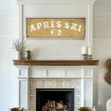 a cozy living room with a fireplace adorned with a decorative tile design. Above the fireplace, there is a framed sign that reads "APRÈS SKI", accompanied by a beer mug icon. The room also includes a wooden shelf with a vase and a woven basket, adding to the rustic charm of the space.