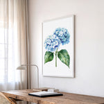 a wooden desk with a lamp, a framed watercolor painting of blue hydrangeas hanging on the wall above it, and a window with sheer curtains in the background.
