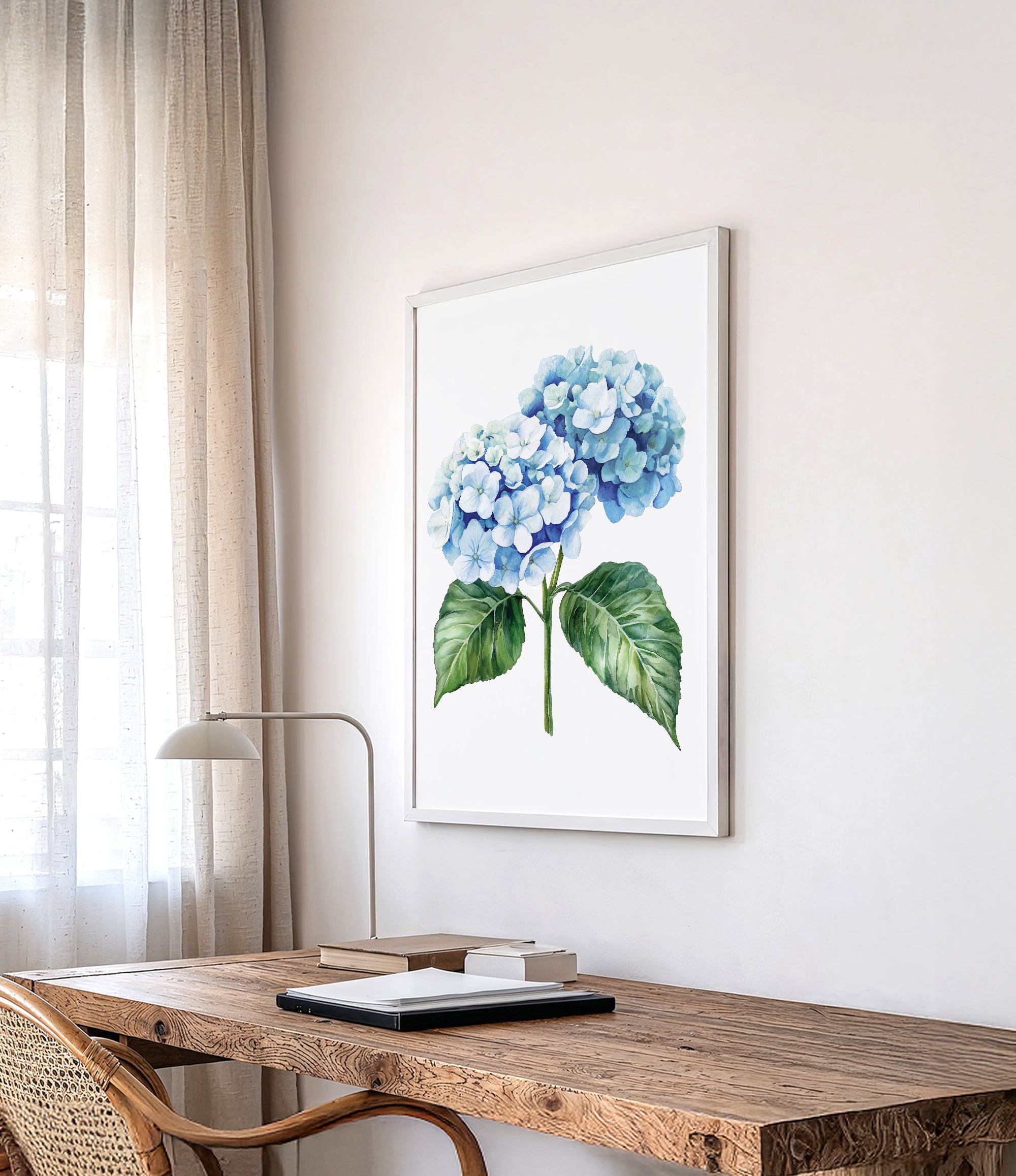 a wooden desk with a lamp, a framed watercolor painting of blue hydrangeas hanging on the wall above it, and a window with sheer curtains in the background.