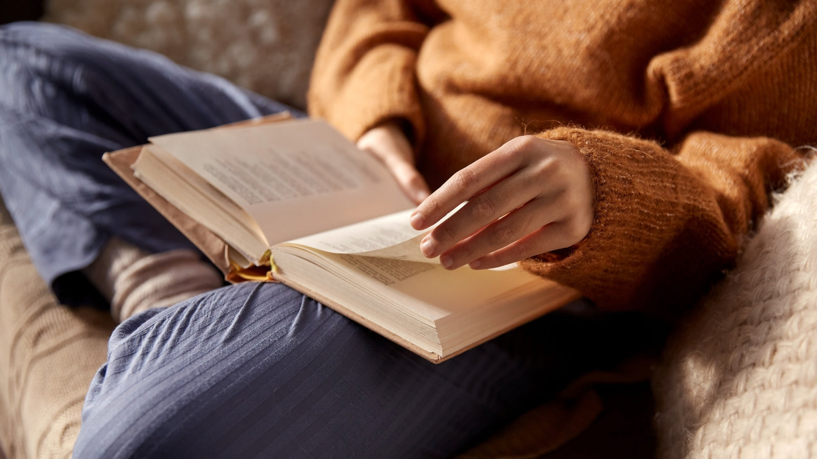 Person wearing a brown sweater and blue pants sitting and reading a book, turning a page.