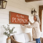 A woman is hanging a framed sign on a wall that says "hey there, PUMPKIN" in a cozy setting with a couch, potted plant, and lantern-style lighting.