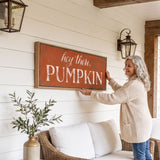 A woman is hanging a framed sign on a wall that says "hey there, PUMPKIN" in a cozy setting with a couch, potted plant, and lantern-style lighting.