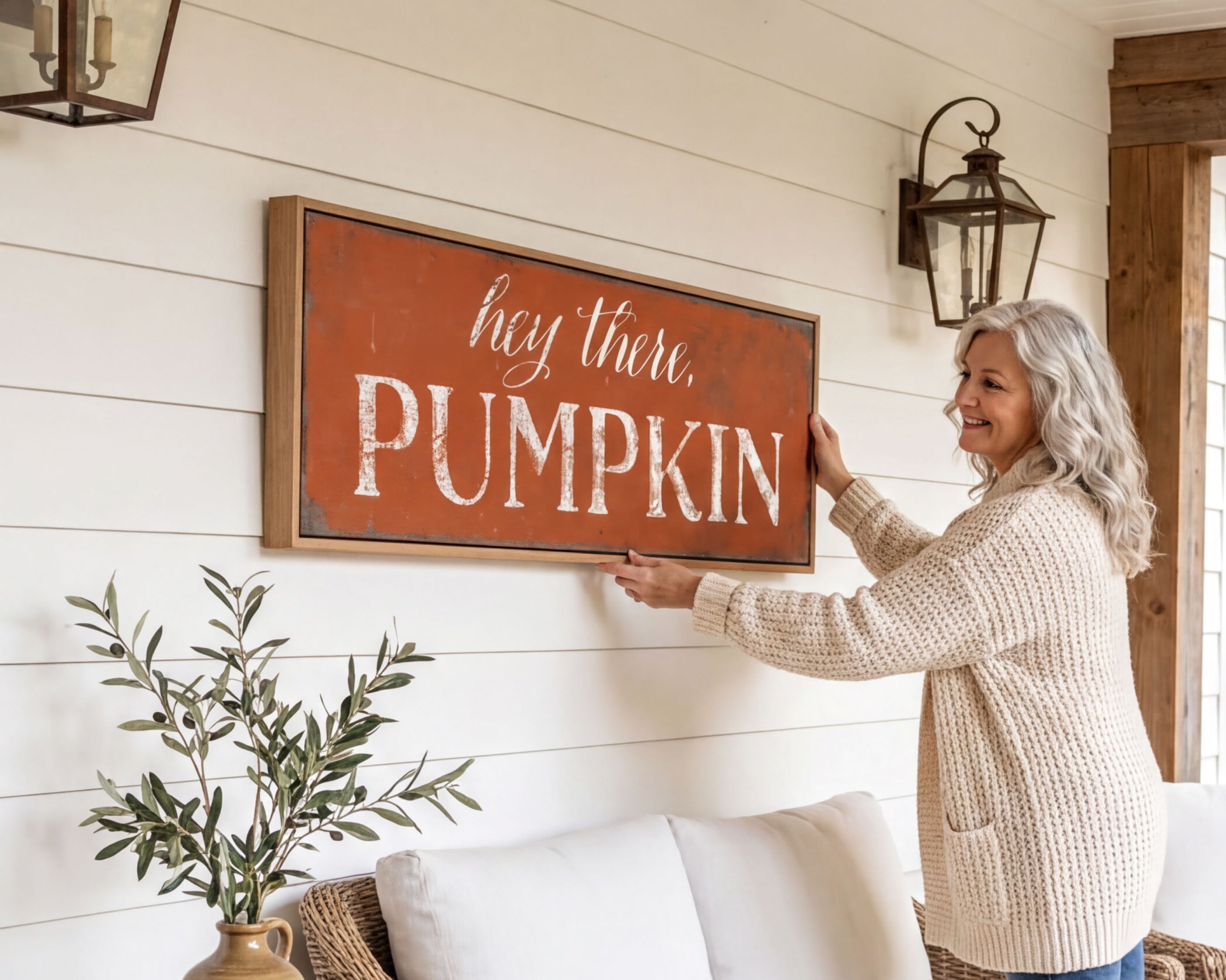 A woman is hanging a framed sign on a wall that says "hey there, PUMPKIN" in a cozy setting with a couch, potted plant, and lantern-style lighting.