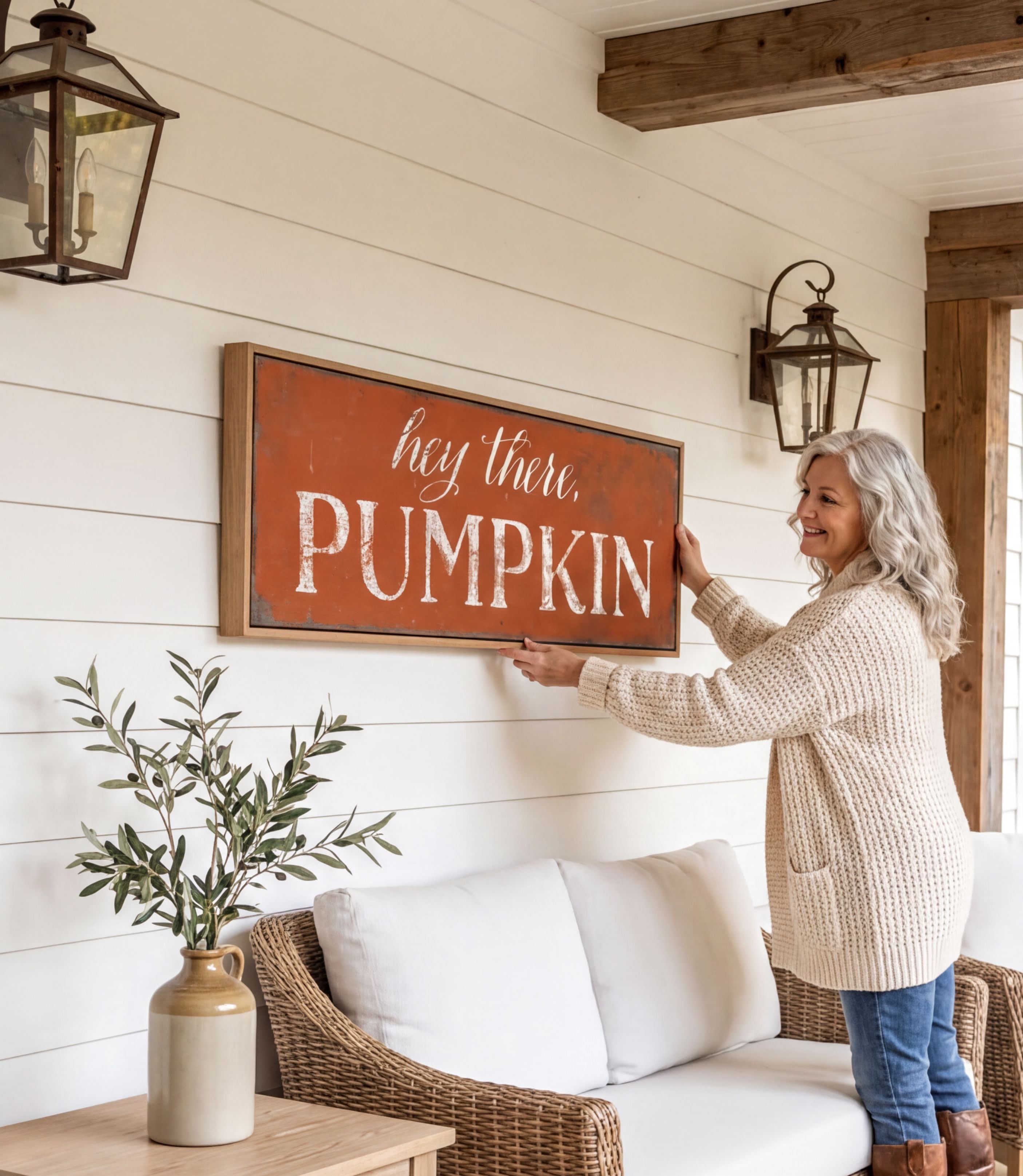 A woman is hanging a framed sign on a wall that says "hey there, PUMPKIN" in a cozy setting with a couch, potted plant, and lantern-style lighting.