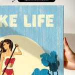 a hand holding a framed print with a beach scene and a woman in a red bikini.