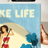 a hand holding a framed print with a beach scene and a woman in a red bikini.