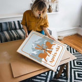 A woman in a yellow shirt is holding a framed picture of a mountain landscape with ski equipment, standing in a room with a patterned rug and a couch.