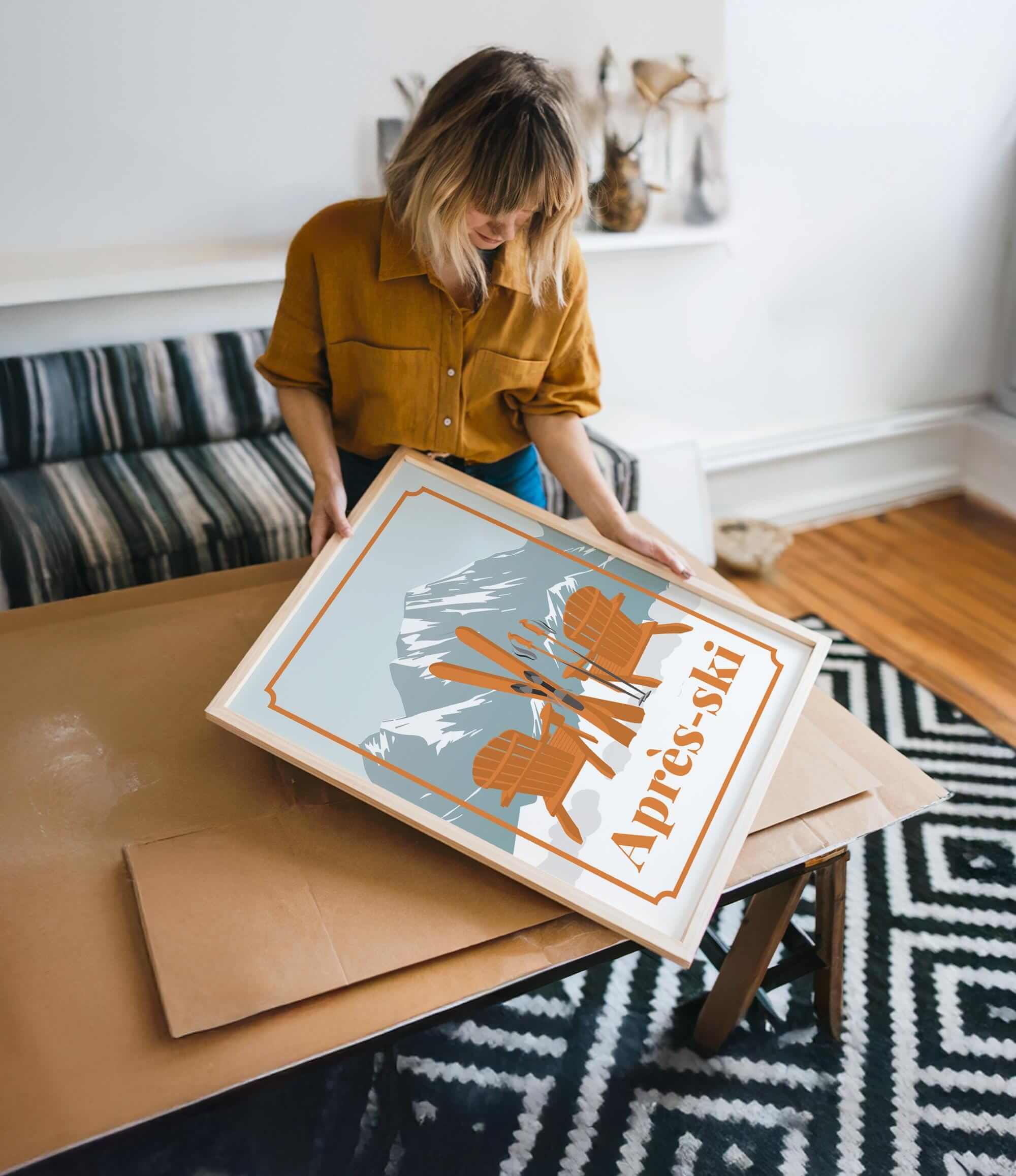 A woman in a yellow shirt is holding a framed picture of a mountain landscape with ski equipment, standing in a room with a patterned rug and a couch.