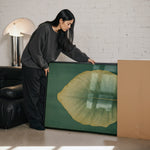 A woman in a gray sweater is leaning over a large framed picture, which features a large green leaf, in a room with a brick wall and a black leather couch.