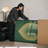 A woman in a gray sweater is leaning over a large framed picture, which features a large green leaf, in a room with a brick wall and a black leather couch.