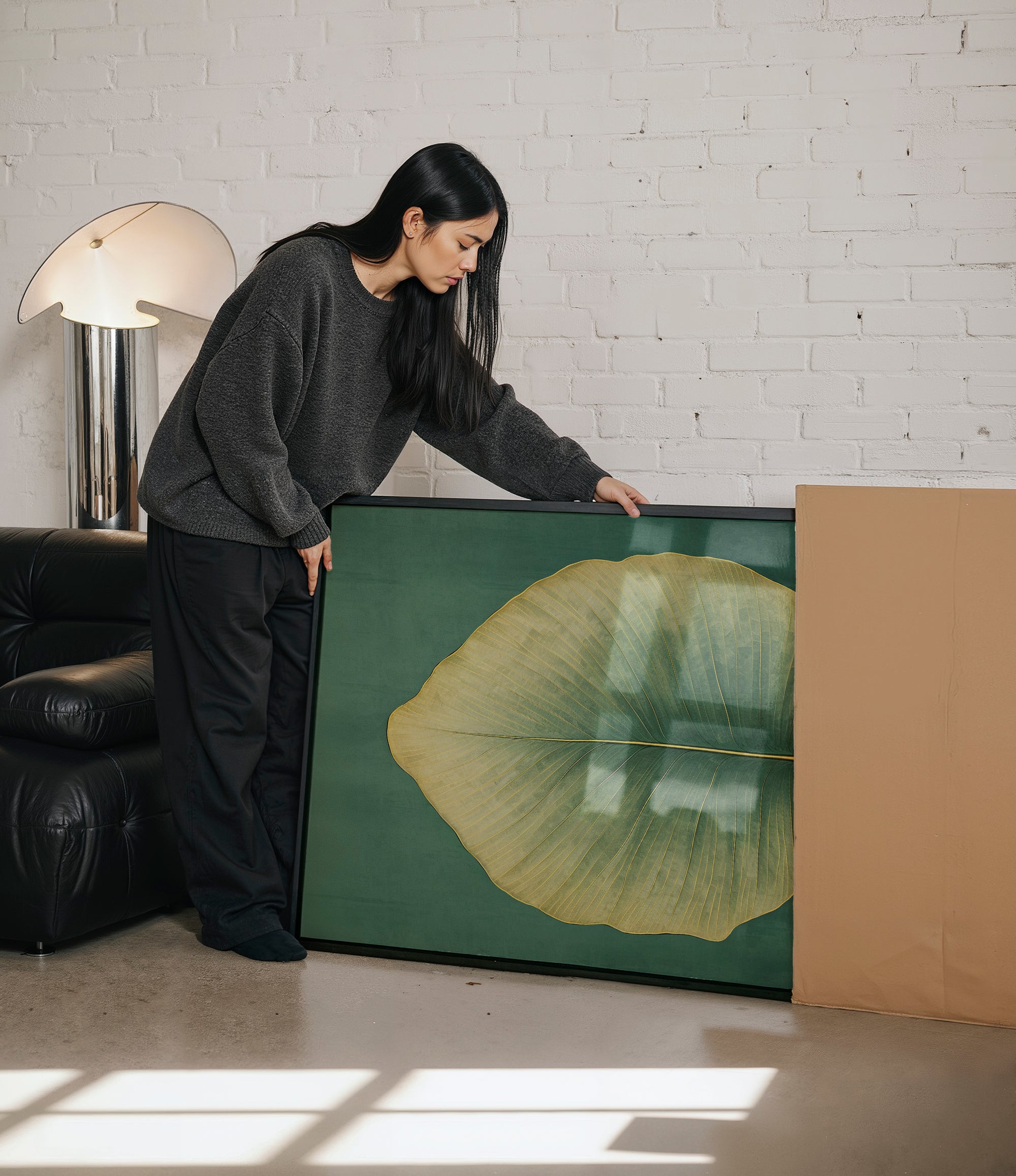 A woman in a gray sweater is leaning over a large framed picture, which features a large green leaf, in a room with a brick wall and a black leather couch.