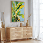 a modern, minimalist interior design with a large, vibrant painting of a banana tree hanging on the wall above a wooden dresser with six drawers. The dresser has a woven vase and a bottle on top, adding a touch of natural elements to the space. The room is bright and airy, with a large window allowing natural light to flood in and a white curtain providing privacy.
