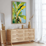 a modern, minimalist interior design with a large, vibrant painting of a banana tree hanging on the wall above a wooden dresser with six drawers. The dresser has a woven vase and a bottle on top, adding a touch of natural elements to the space. The room is bright and airy, with a large window allowing natural light to flood in and a white curtain providing privacy.