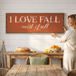 A woman is standing in front of a rustic wooden table, holding a large sign that reads "I LOVE FALL most of all". The table has a loaf of bread, a bowl of oranges, and a wicker chair.