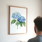 A framed watercolor painting of blue hydrangeas hangs on a white wall, with a person standing in the foreground looking at the artwork.