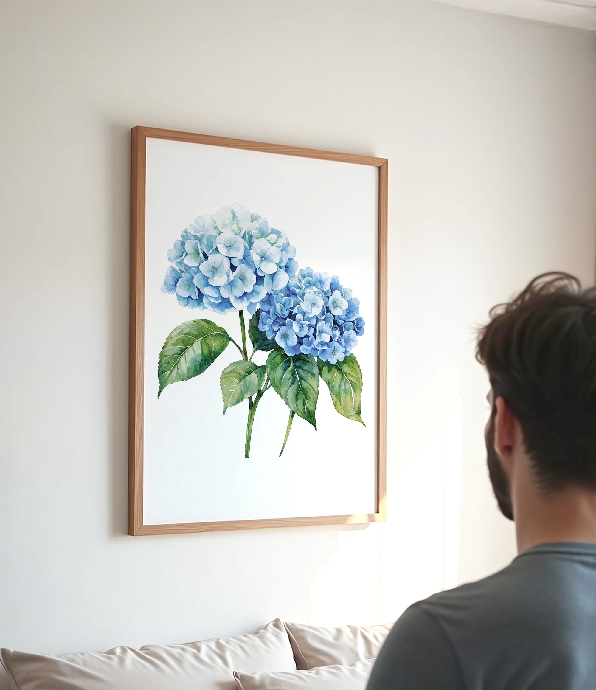 A framed watercolor painting of blue hydrangeas hangs on a white wall, with a person standing in the foreground looking at the artwork.