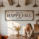 A woman is sitting at a wooden table in a cozy kitchen, holding a cup and smiling. Above her, a rustic sign reads "Have a HAPPY FALL y'all". The table is set with a vase of autumn leaves and a plate of food, creating a warm and inviting atmosphere.