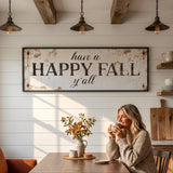 A woman is sitting at a wooden table in a cozy kitchen, holding a cup and smiling. Above her, a rustic sign reads "Have a HAPPY FALL y'all". The table is set with a vase of autumn leaves and a plate of food, creating a warm and inviting atmosphere.