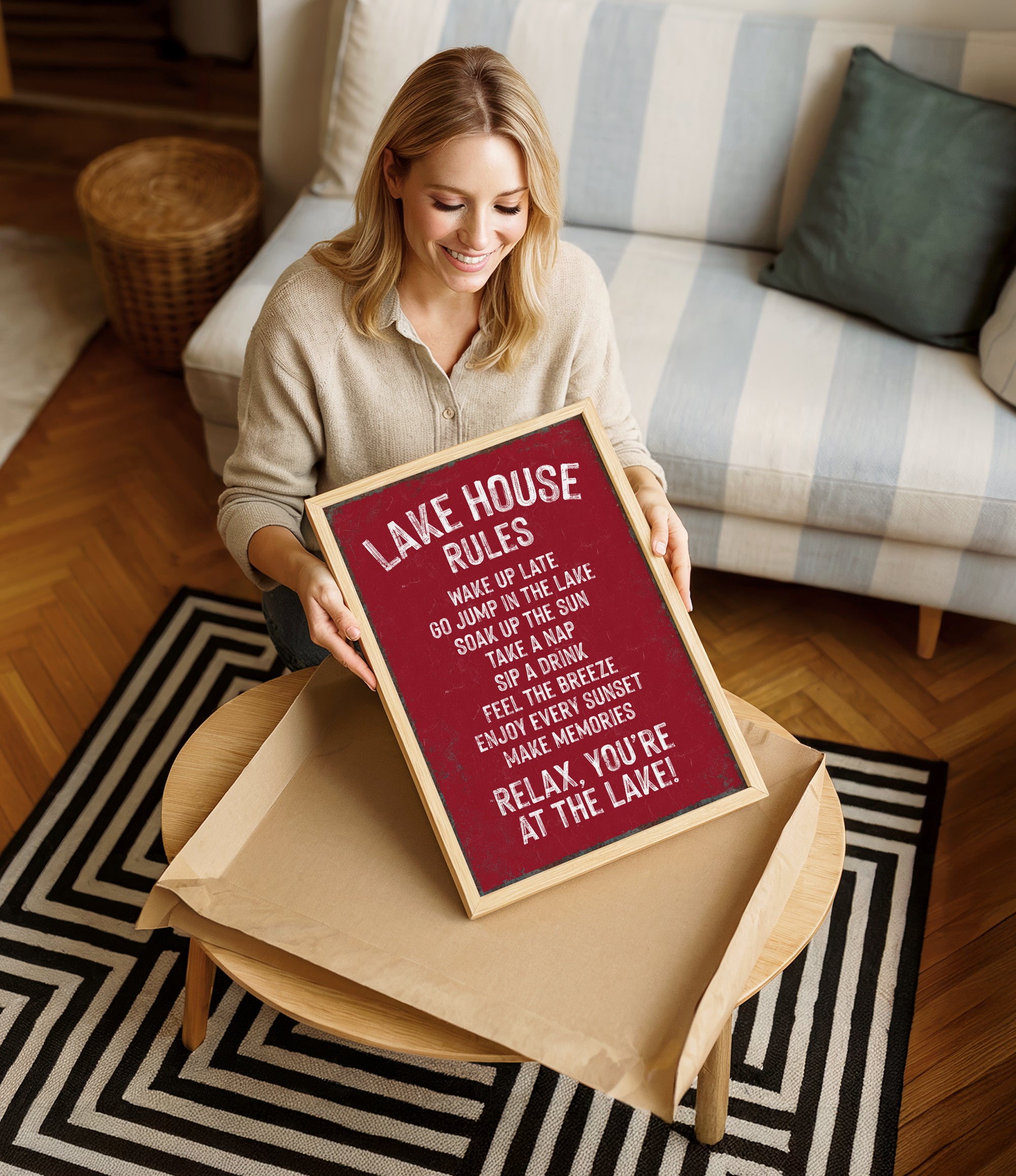A woman is sitting on a couch and holding up a framed sign with a red background and white text.