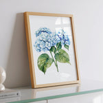 A framed watercolor painting of blue hydrangea flowers is displayed on a white dresser, with a book and a lamp visible in the background.