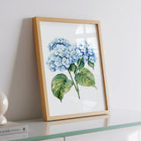 A framed watercolor painting of blue hydrangea flowers is displayed on a white dresser, with a book and a lamp visible in the background.