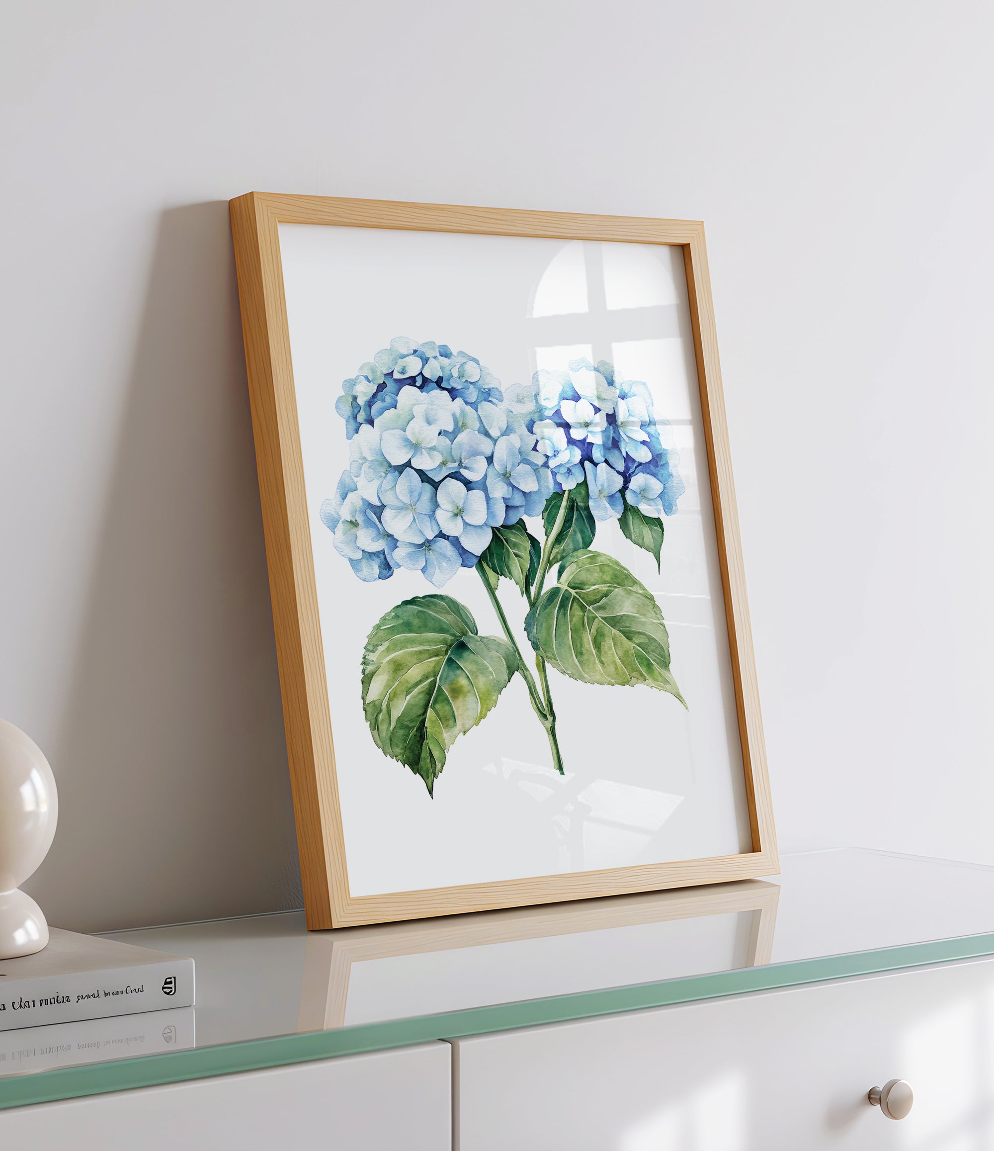 A framed watercolor painting of blue hydrangea flowers is displayed on a white dresser, with a book and a lamp visible in the background.