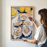 A woman is hanging a framed artwork on a wall, which features a vibrant painting of oysters and lemons.