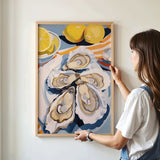 A woman is hanging a framed artwork on a wall, which features a vibrant painting of oysters and lemons.