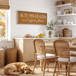 A cozy kitchen scene with a dog resting on the floor, a wooden table and chairs, and a rustic wall sign that reads "Autumn Leaves and apples please".