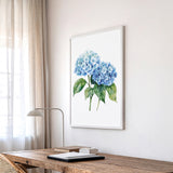 a wooden desk with a lamp, a framed watercolor painting of blue hydrangeas hanging on the wall above it, and a window with sheer curtains in the background.