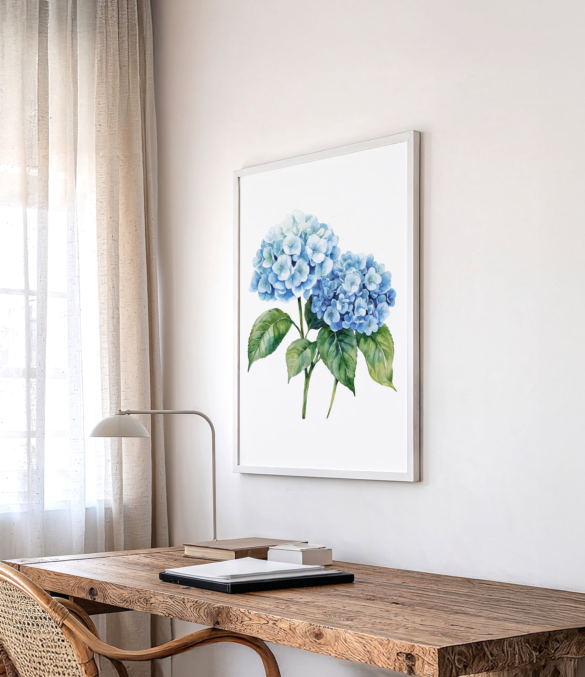 a wooden desk with a lamp, a framed watercolor painting of blue hydrangeas hanging on the wall above it, and a window with sheer curtains in the background.