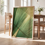 a large framed print of a green leaf with a yellow center, displayed on a wooden floor in a room with a dining table and chairs.